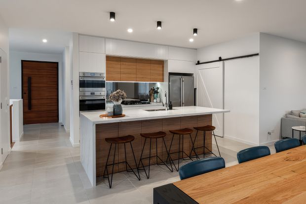 Bright contemporary open-plan kitchen and dining area. The kitchen features a large white island with an integrated sink and seating for four, white full-height cabinetry, warm timber cabinetry accents, stainless-steel appliances, pale tiled flooring, and black track-style pendant spotlights. A timber front door and hallway are visible to the left, while a dining table and blue upholstered chairs occupy the foreground.