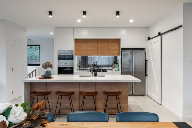 A bright contemporary kitchen with a large island and four bar stools, white perimeter cabinetry, warm timber drawer fronts and overhead doors, a white stone-look benchtop, integrated appliances, stainless-steel refrigerator, and a concealed barn-style sliding door. A dining table and blue dining chairs are visible in the foreground, with a hallway and stair area beyond.