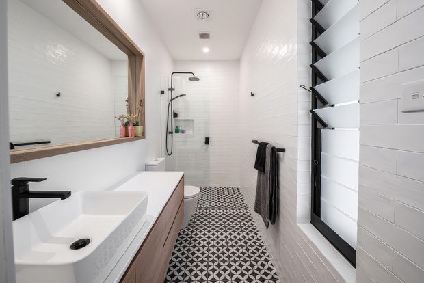 Bright contemporary bathroom with a long white vanity, rectangular vessel basin and matte black tapware. White tiled walls, a frameless glass shower, black geometric patterned floor tiles, timber-framed mirror, black-framed louvred window, and dark towel rails create a crisp monochrome scheme with warm timber accents.