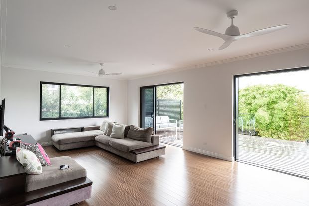 Bright, spacious contemporary living room with pale timber flooring, neutral modular sofas, large black-framed windows and sliding glass doors opening to a timber deck. The room features a white ceiling with recessed lighting, decorative cornices and two ceiling fans, with leafy greenery visible outside.