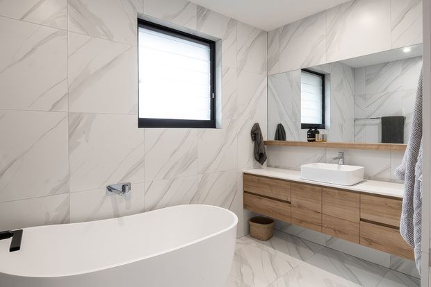 Contemporary bathroom featuring full-height white marble-look wall and floor tiles with grey veining, a freestanding white bathtub, floating timber vanity with a white vessel basin, large mirror, black-framed window, and understated black fixtures.