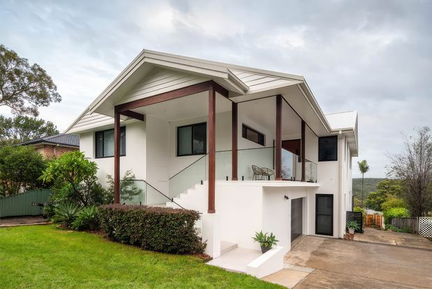 Exterior view of a contemporary split-level residential house with a prominent white gabled roof, white rendered walls, dark timber posts, glass balustrades, and black-framed windows and doors. A raised terrace and staircase sit above a lower-level entry or garage area, with landscaped garden and concrete driveway in front.