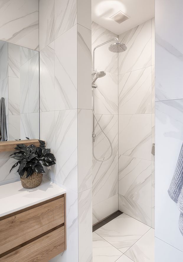 A bright contemporary bathroom featuring a walk-in tiled shower with large white marble-look porcelain tiles and fine grey veining. The shower has chrome rain and handheld fixtures, a clear glass screen, and a dark linear floor drain. A timber-veneer vanity with a white benchtop and a dark leafy potted plant sits beside the shower, with a wall mirror above.