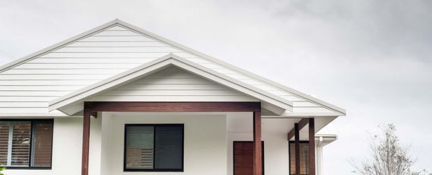 Exterior view of a low-rise house with overlapping gabled roof forms, white horizontal weatherboard cladding, dark-framed windows, and a timber-clad recessed entry porch supported by dark timber posts. The facade has a clean contemporary residential appearance under an overcast sky.