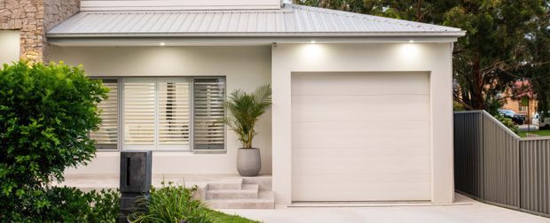 Street-facing view of a compact contemporary residential facade with a pale rendered exterior, white metal low-pitched roof, integrated single garage door, shuttered window, small entry stair and landscaped planting. A stone-clad wall is partially visible at the left edge, with a side boundary fence and driveway to the right.