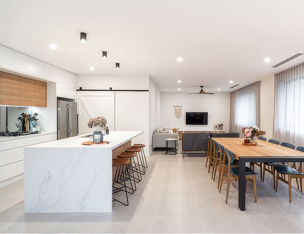 Bright open-plan contemporary kitchen, dining, and living area. The kitchen features white flat-panel cabinetry, warm timber overhead cupboards, a large white veined stone island with bar seating, and integrated stainless-steel appliances. A timber dining table sits beside the living area, which includes a grey sofa, wall-mounted television, and large sheer-curtained windows. Recessed and pendant lighting create a clean, airy interior.
