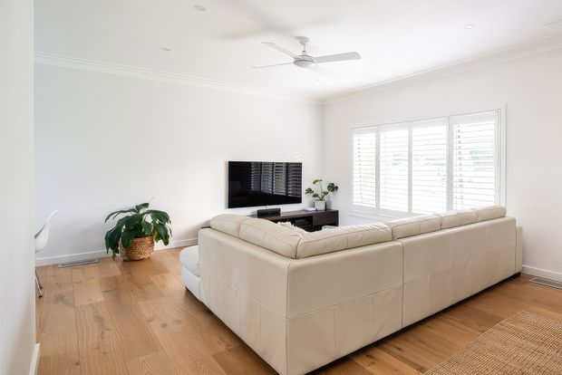 Bright, spacious living room with white walls, decorative ceiling cornices, a white ceiling fan, timber flooring, plantation shutters, and a large cream sectional sofa facing a wall-mounted television. Potted plants and a woven rug add natural texture.