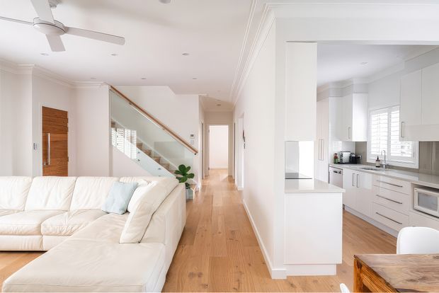 Bright open-plan residential interior showing a living room in the foreground, a contemporary white kitchen to the right, and a hallway with a timber staircase and glass balustrade leading toward the entry. The space features pale timber flooring, white walls and cabinetry, timber door detailing, plantation shutters, recessed lighting, and a ceiling fan.