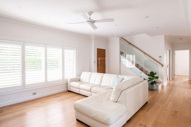 Bright contemporary living room with a large white sectional sofa, pale timber flooring, white plantation shutters, recessed ceiling lights, and a ceiling fan. A timber front door and a glass-panelled staircase are visible, with a hallway extending beyond the room. The space has a clean, airy, neutral presentation with warm wood accents.