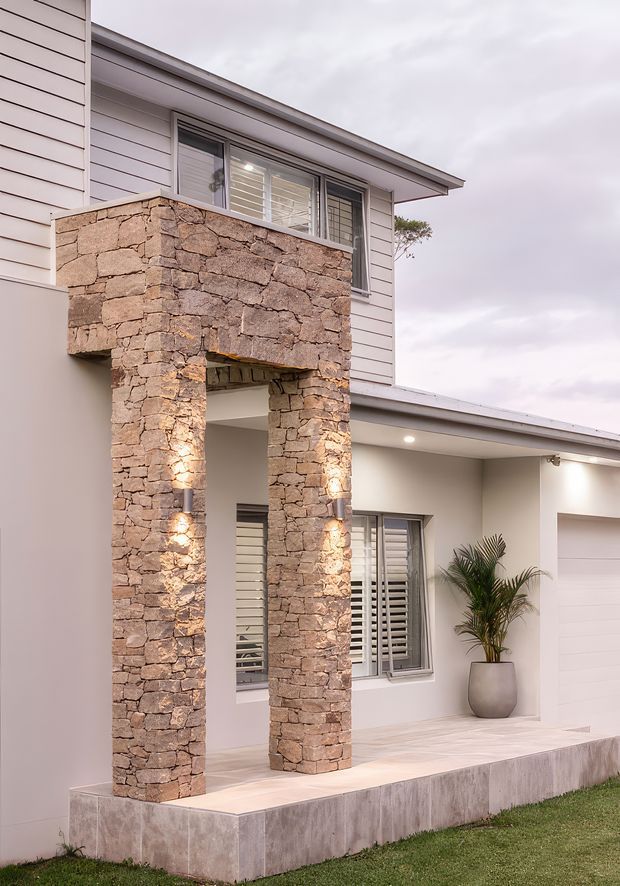 Exterior view of a contemporary two-storey home entrance, featuring pale horizontal weatherboard cladding, a projecting stone-clad portal with square columns, sheltered porch, plantation-shuttered windows, recessed lighting, a potted palm, and an integrated garage.
