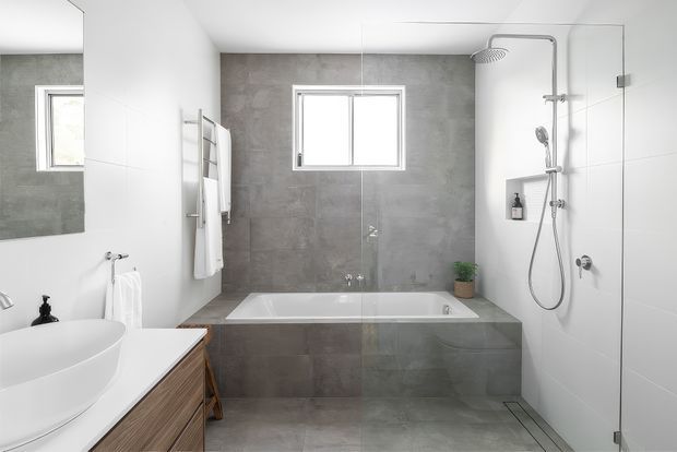 Bright contemporary bathroom with a freestanding white basin on timber vanity cabinetry, a built-in bathtub set into grey concrete-look tiled walls, frameless glass shower enclosure with overhead and handheld shower fittings, large mirror, and white wall surfaces.