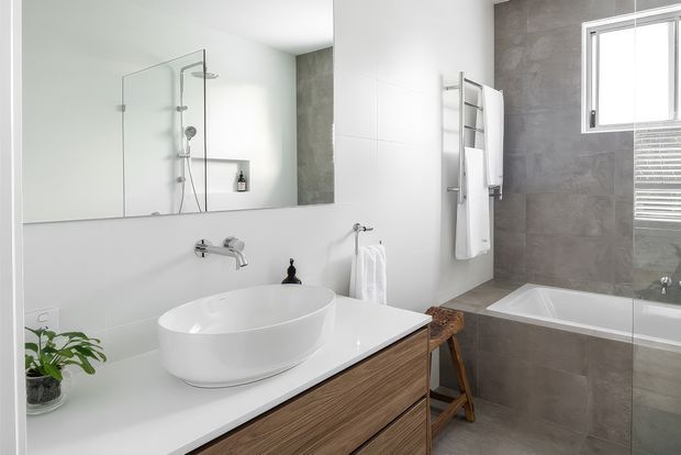 Bright contemporary bathroom featuring a floating timber vanity with a white vessel basin, wall-mounted tapware, a frameless glass shower, freestanding shower fixtures, and a tiled bathtub surround. Grey stone-look wall and bath tiles contrast with white surfaces, while a window provides natural light.