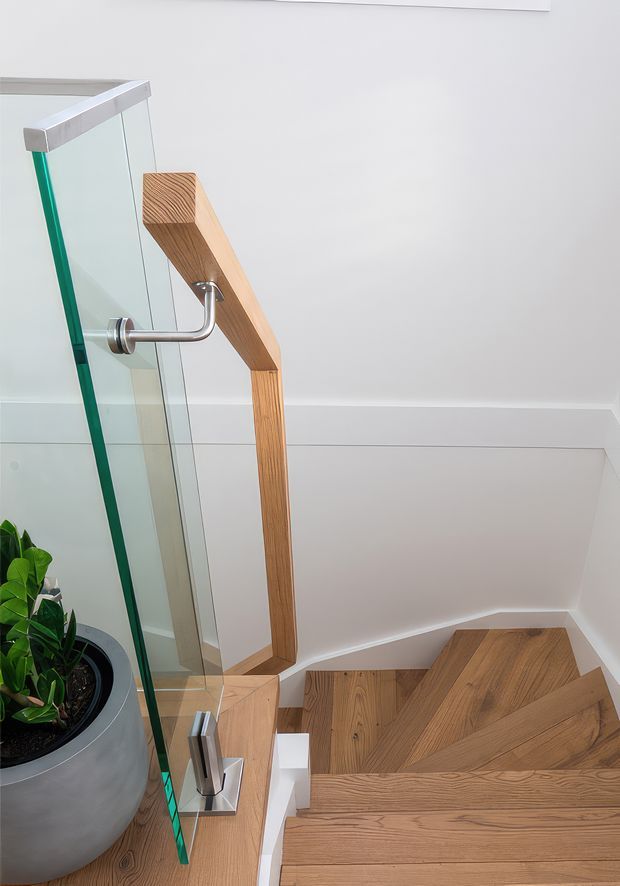 A close-up interior view of a contemporary stairway with warm timber treads and a matching timber handrail, paired with clear glass balustrading and brushed metal fittings. White walls and trim create a clean, bright setting, with a potted plant beside the stairs.