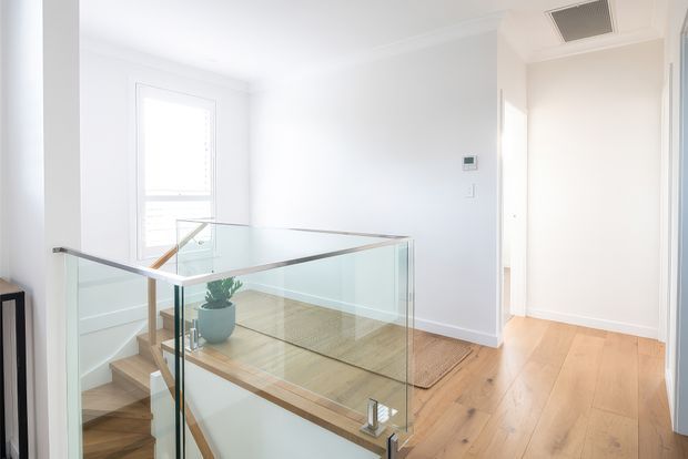 Bright upper-level landing and stairwell in a contemporary home, featuring pale timber flooring, white walls and trim, and a clear glass balustrade with timber handrail. A stair descends beside a window, while a hallway and ceiling air vent are visible to the right.
