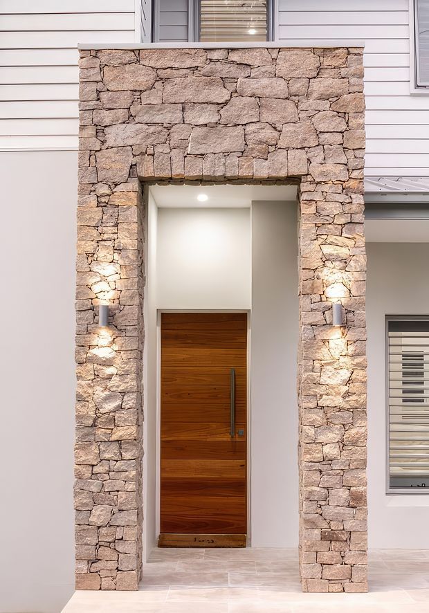 Front exterior entry of a contemporary home, featuring a tall recessed timber door framed by a substantial rough natural-stone portal. White horizontal weatherboard cladding, simple rectangular windows with blinds, pale tiled paving, and paired wall sconces complete the facade.