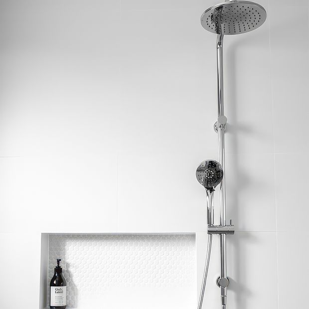 Close-up of a contemporary bathroom shower installation. A chrome rail-mounted shower system features a large round overhead shower, handheld shower, controls and hose against white tiled walls. A recessed wall niche with small white textured tiles holds a dark soap dispenser.