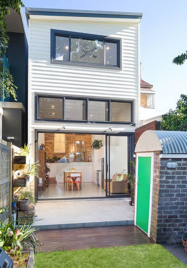 Exterior view of a narrow multi-storey urban dwelling with a white horizontal weatherboard facade, dark-framed windows, and large glazed folding or sliding doors opening onto a compact timber deck and lawn. The ground level reveals a bright open-plan kitchen and living area with white cabinetry, pale flooring, exposed brick detailing, and a small dining table. A curved corrugated-roof brick outbuilding with a vivid green door occupies the right side of the courtyard.
