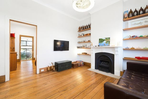 Bright traditional living room with polished honey-toned timber floorboards, white walls and ceiling, built-in open display shelving, a decorative black fireplace with white mantel, wall-mounted television, leather sofa, and a doorway opening to another timber-floored room. Bottles and small objects are displayed on the shelves.
