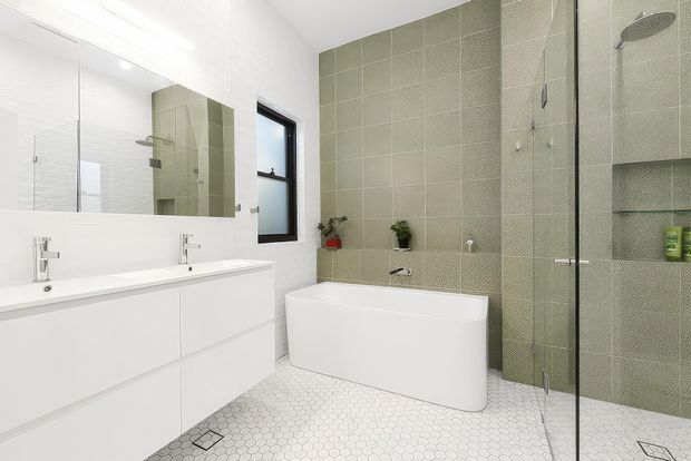 Bright contemporary bathroom with a freestanding white bathtub, double white vanity, large mirror, glass shower enclosure, and green textured wall tiles. The floor is finished in small white hexagonal tiles, with black-framed window details and minimal plant accents.