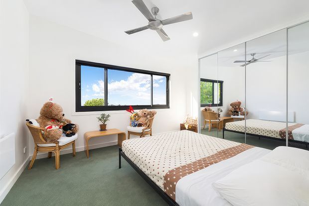 Bright, playful bedroom with a large bed, green carpet, white walls, black-framed window, mirrored built-in wardrobes, ceiling fan, and a small seating area with teddy bears and a side table. The room has abundant daylight and a cheerful, child-friendly character.