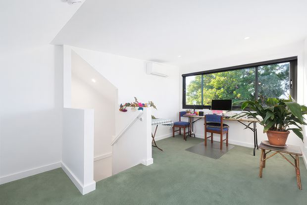 Bright, minimalist home office on an upper landing with pale green carpet, white walls and ceiling, a large black-framed window overlooking trees, and a long desk with computer and chairs. A stair opening with a simple white balustrade connects to the lower level, while indoor plants and a small floral arrangement add natural accents.