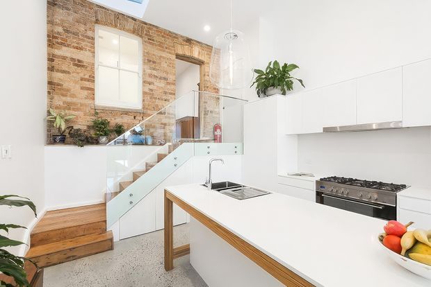 Bright contemporary kitchen with a large white island featuring timber edging, integrated sink, and a stainless-steel range cooker. White flat-panel cabinetry spans the right wall, while a glass balustrade encloses stairs leading to a mezzanine or upper level. Exposed brick forms a warm feature wall with a small window and greenery, contrasting with the polished concrete or terrazzo-like floor and white walls.