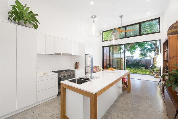 Bright contemporary kitchen with white full-height cabinetry, a large white island with timber edging and integrated sink, stainless steel cooking appliances, polished concrete flooring, pendant lights, and expansive glazing opening to a leafy outdoor area.