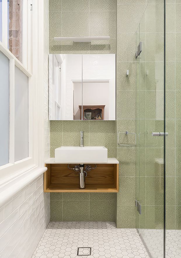 Bright compact bathroom with a wall-mounted timber vanity, white rectangular vessel basin, chrome tapware, large mirror, and frameless glass shower enclosure. The walls and shower are clad in pale sage-green textured tiles, while the floor uses small white hexagonal tiles. A tall window provides abundant natural light, and a reflected fireplace is visible in the mirror.