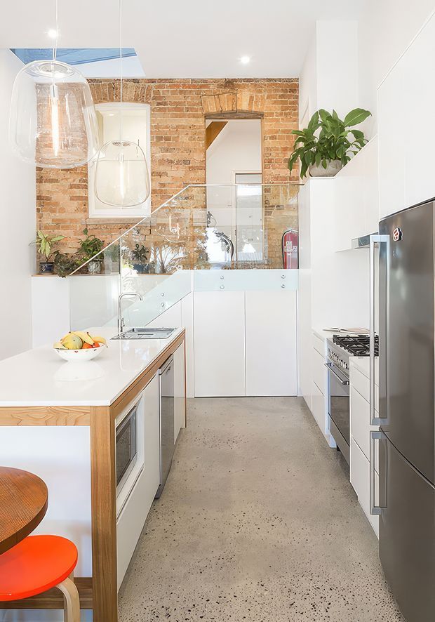 Bright, narrow galley-style kitchen with white full-height cabinetry, white benchtops, stainless-steel appliances, a polished concrete floor, and an exposed red-brick feature wall. A timber dining peninsula with colourful stools occupies the foreground. Glass pendant lights, mirrored splashback panels, indoor plants, and a skylight add brightness and visual openness.