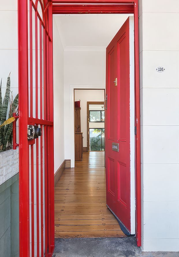 A residential entrance viewed from outside, featuring a vivid red metal security gate and matching red paneled front door. The doorway opens into a narrow hall with warm polished timber floorboards, white walls, timber trim, and a glimpse of a timber-furnished room and rear glazing beyond. A house number plaque is visible on the exterior wall.