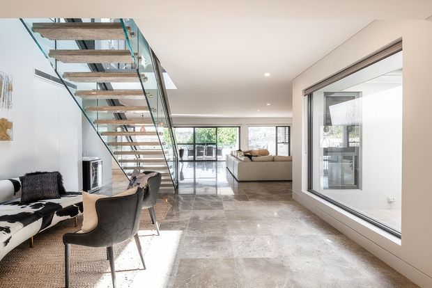 Bright contemporary open-plan interior with a polished stone or concrete-look floor, white walls, expansive glazing, and a floating staircase with thick timber treads and clear glass balustrades. A living area with a neutral sofa is visible at the rear, while a cowhide-upholstered seating area and dining chairs occupy the foreground. A large internal glazed opening provides a view into an adjacent room.