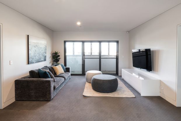 A bright, contemporary living room with a grey sectional sofa, two rounded ottomans, a light rug, wall-mounted television, and sleek white media cabinetry. Large glazed doors or windows provide natural light, while neutral finishes and minimal furnishings create an airy, understated interior.