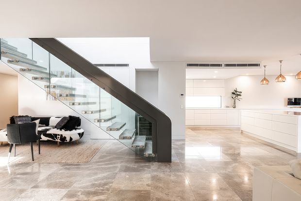 Bright open-plan contemporary interior showing a minimalist white kitchen and living area, with a sculptural floating staircase featuring a dark structural side rail and clear glass balustrade. The space has highly polished large-format stone or marble-look flooring, integrated white cabinetry, pendant lights, and a small seating area.