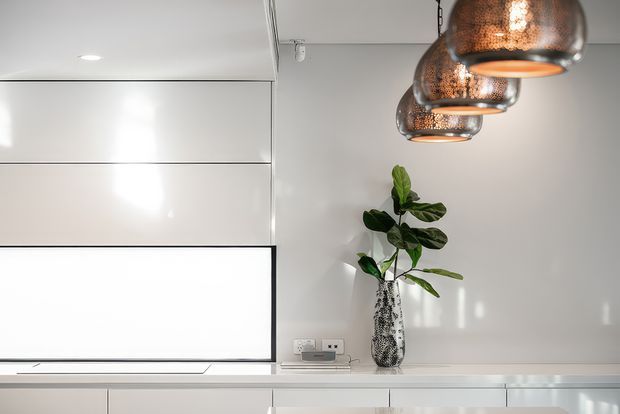 A clean, minimalist interior wall featuring glossy white built-in cabinetry or media joinery, a wall-mounted black-framed screen, a slim white console, and a potted plant in a textured vase. A row of three copper pendant lights hangs from the ceiling, adding warmth to the otherwise white scheme.