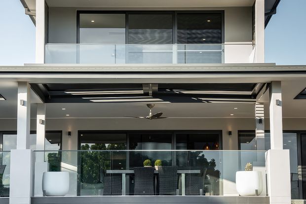 Exterior view of a contemporary multi-level residential building with a covered outdoor entertaining area, glass balustrades, dark-framed glazing, white rendered walls, and a modern pergola or retractable roof structure. The lower level features outdoor dining furniture and large planters, while an upper balcony is visible above.