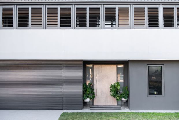 Front exterior of a contemporary multi-level residence with a clean white and grey facade. The upper level features a continuous row of louvred windows, while the ground floor includes a wide horizontal roller garage door, a recessed timber-toned entry door with potted plants, and a narrow vertical window. A small strip of lawn borders the facade.