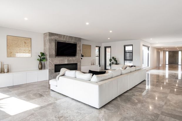Bright, spacious contemporary living room with a large white sectional sofa, polished stone-look tiled flooring, and a feature wall incorporating a wall-mounted television and fireplace. White built-in cabinetry, neutral artwork, indoor plants, recessed ceiling lights, and extensive glazing create an open, refined interior extending toward an entry or hallway.