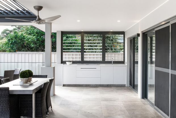 A bright covered outdoor entertaining area with a built-in white kitchenette and extensive dark-framed louvred glazing. A dining table with woven chairs occupies the foreground, while a ceiling fan, tiled floor, white privacy fence and lush greenery create a relaxed indoor-outdoor setting.