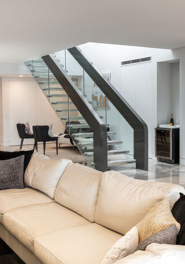 Bright contemporary living room with a large cream sectional sofa in the foreground, a glass-and-metal sculptural staircase, polished stone flooring, integrated wall cabinetry, and a compact built-in bar or wine cabinet. A dining area and artwork are visible in the background.
