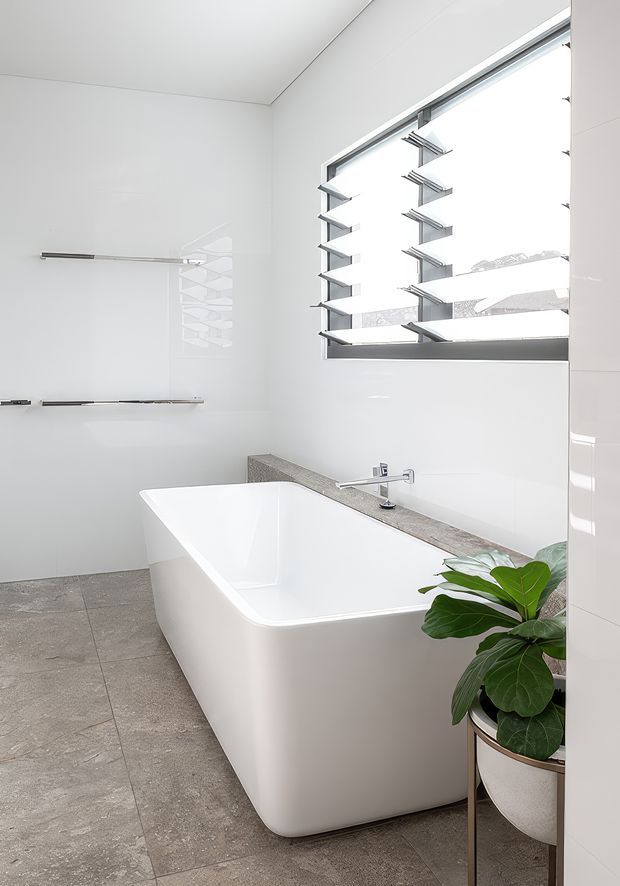 Bright contemporary bathroom featuring a freestanding white bathtub, large grey stone-look floor tiles, white wall surfaces, a slim grey benchtop ledge with wall-mounted tapware, and a wide aluminium louvred window. A potted leafy plant adds a natural accent.