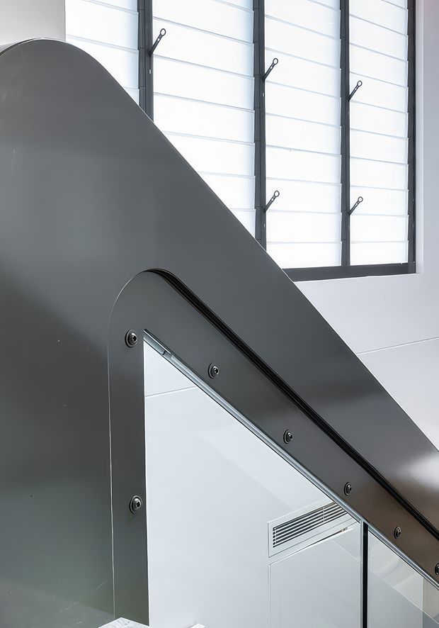 Close-up architectural interior detail showing a dark grey metal stair or ramp balustrade with rounded edges and exposed fasteners, positioned diagonally across the frame. Behind it are tall translucent louvred or panelled windows with dark metal framing, white walls, and a reflected view of a bright interior with a linear air vent. The image has a clean, industrial-modern character.