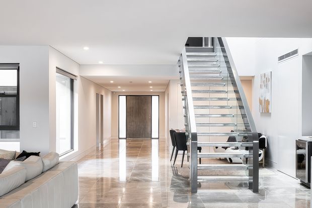 Bright, spacious contemporary open-plan interior featuring a polished stone floor, white walls and ceiling, expansive glazing, and a prominent floating staircase with glass balustrades and dark metal supports. A living area is visible at left, with a dining area beneath the stair and a hallway extending toward a dark feature door.