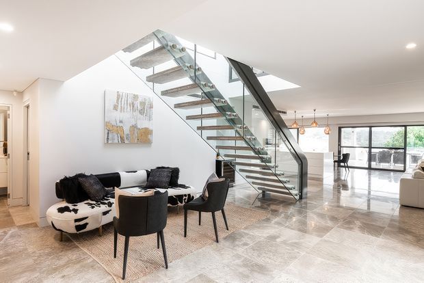Bright open-plan contemporary interior featuring a floating staircase with timber treads, glass balustrades and a dark structural stringer. The foreground contains a seating area with a sofa, armchairs, cowhide ottoman and rug, while a dining and kitchen area is visible beyond. Polished stone flooring, white walls, skylights and extensive glazing create an airy, refined space.