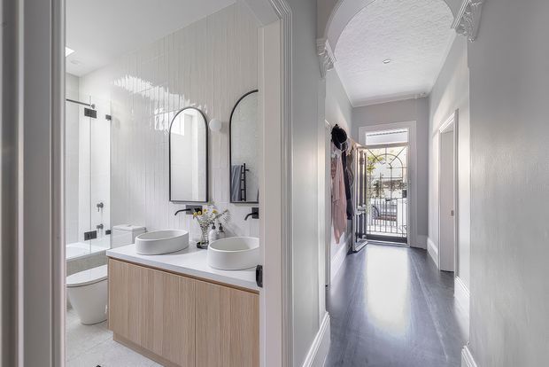 A bright, refined interior showing a contemporary bathroom opening directly onto a long hallway. The bathroom features a double vanity with vessel basins, light timber-look cabinetry, white vertically textured wall tiles, arched mirrors, and a toilet and shower. The hallway has dark timber flooring, white walls, decorative arched ceiling detailing, and a glazed security entry door with sidelights.