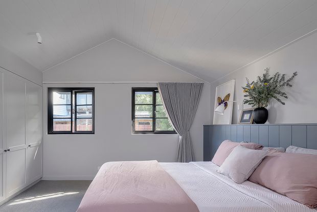 Bright contemporary bedroom with a pitched, panelled ceiling, soft grey walls, built-in white wardrobes, black-framed windows, and a large bed dressed in blush pink and white bedding. A pleated grey curtain, blue-grey headboard wall, framed artwork, and a dark decorative vase add subtle colour and texture.