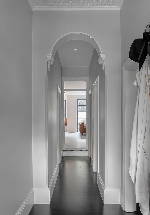 A narrow interior hallway viewed toward a brighter living area, with dark flooring, light grey walls, white trim and baseboards, multiple doorways, and a decorative arched opening with classical moulded detailing. Coats and hats hang at the right edge, suggesting an entry or circulation area. The corridor has standard ceiling height and a calm, refined traditional character.
