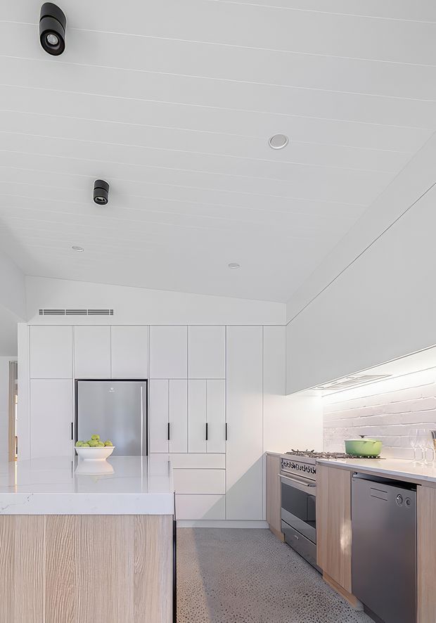 Bright contemporary kitchen with white full-height cabinetry, a large island with a pale timber-look base and white benchtop, stainless-steel appliances, and a secondary timber-toned island run. White painted brick forms the splashback wall, with integrated under-cabinet lighting and a polished concrete or terrazzo-like floor. The ceiling has recessed lighting and directional black spotlights.