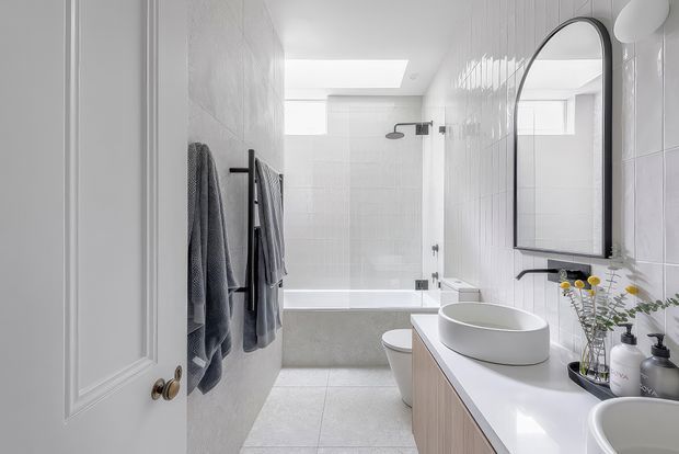 Bright contemporary bathroom viewed from the doorway, featuring white tiled walls, a skylight, bathtub with shower, wall-mounted towel rails, a long white vanity with timber cabinetry, vessel basins, and a large arched black-framed mirror.