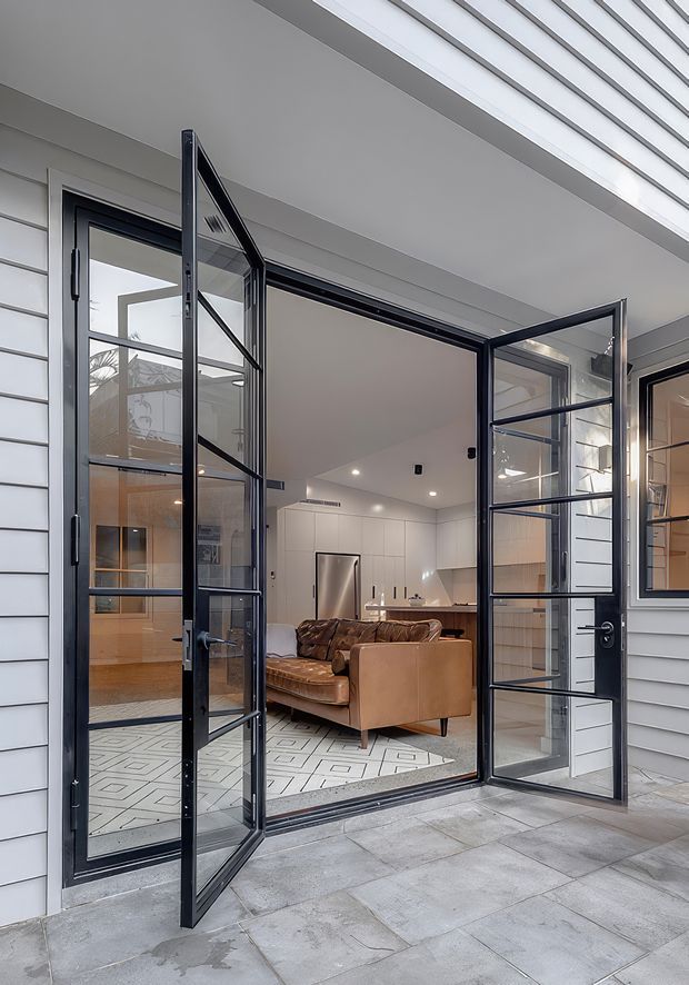 View from a covered outdoor patio through open black-framed glazed bi-fold doors into a contemporary open-plan living and kitchen area. The exterior has white horizontal weatherboard cladding and grey tiled paving. Inside, a tan leather sofa sits on a patterned rug, with white built-in kitchen cabinetry, stainless-steel appliances, recessed lighting, and warm timber details visible beyond.