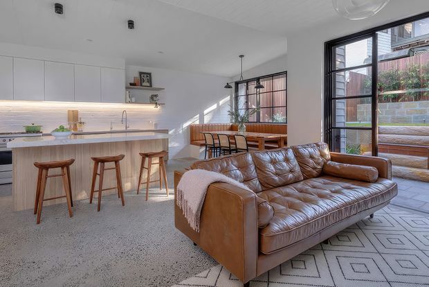 Bright open-plan living area combining a modern kitchen, dining zone, and lounge. The space features white flat-panel cabinetry, a pale stone or composite island benchtop, integrated warm under-cabinet lighting, terrazzo-style flooring, large black-framed glazed doors and windows, a timber dining table, and a prominent brown tufted leather sofa over a geometric rug. A paved outdoor courtyard is visible through the glazing.